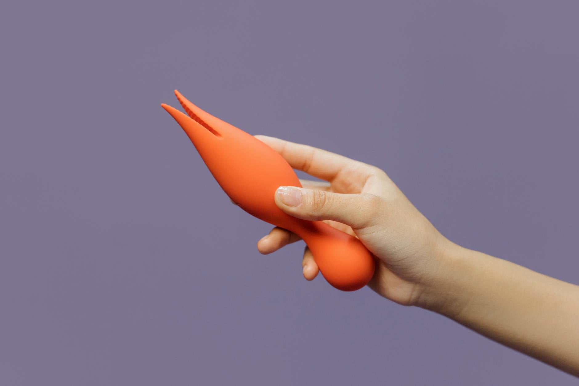 Eine Hand hält einen orangefarbenen Lemon Vibrator vor minimalistischem lila Hintergrund, modernes Schönheitsgefühl