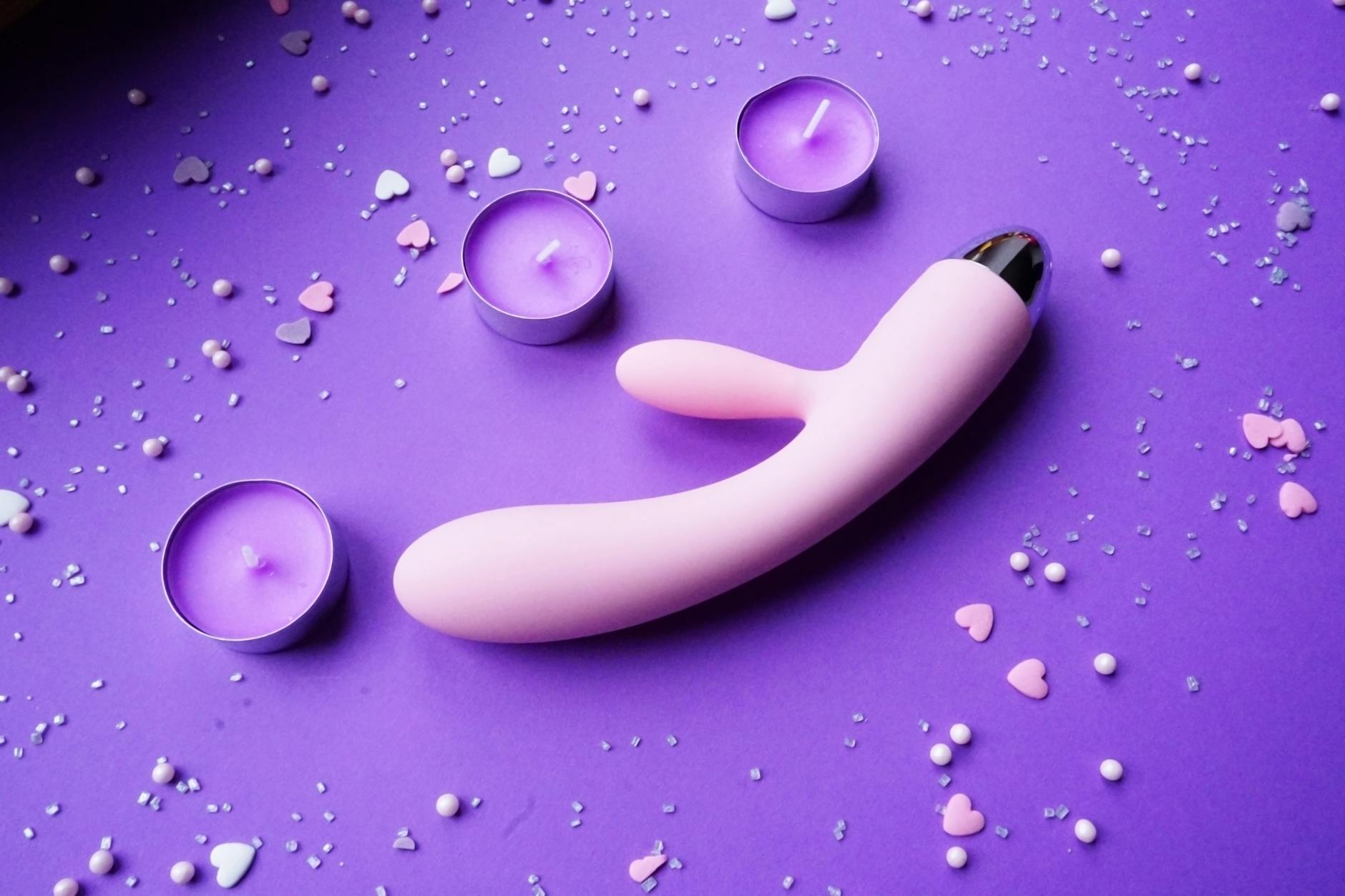 Ein rosa Saugvibrator auf violettem Hintergrund mit Herzchen-Konfetti und Kerzen für eine romantische Atmosphäre